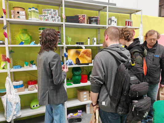 Visitors gather around a 3D printing display booth showcasing colorful filament samples, finished prints, and a screen playing a demo of the Hanboost Ultrasonic Cutter in use.